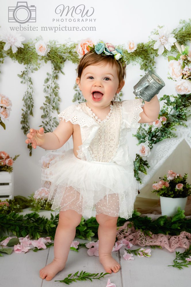 A baby girl in white dress holding up a cell phone.