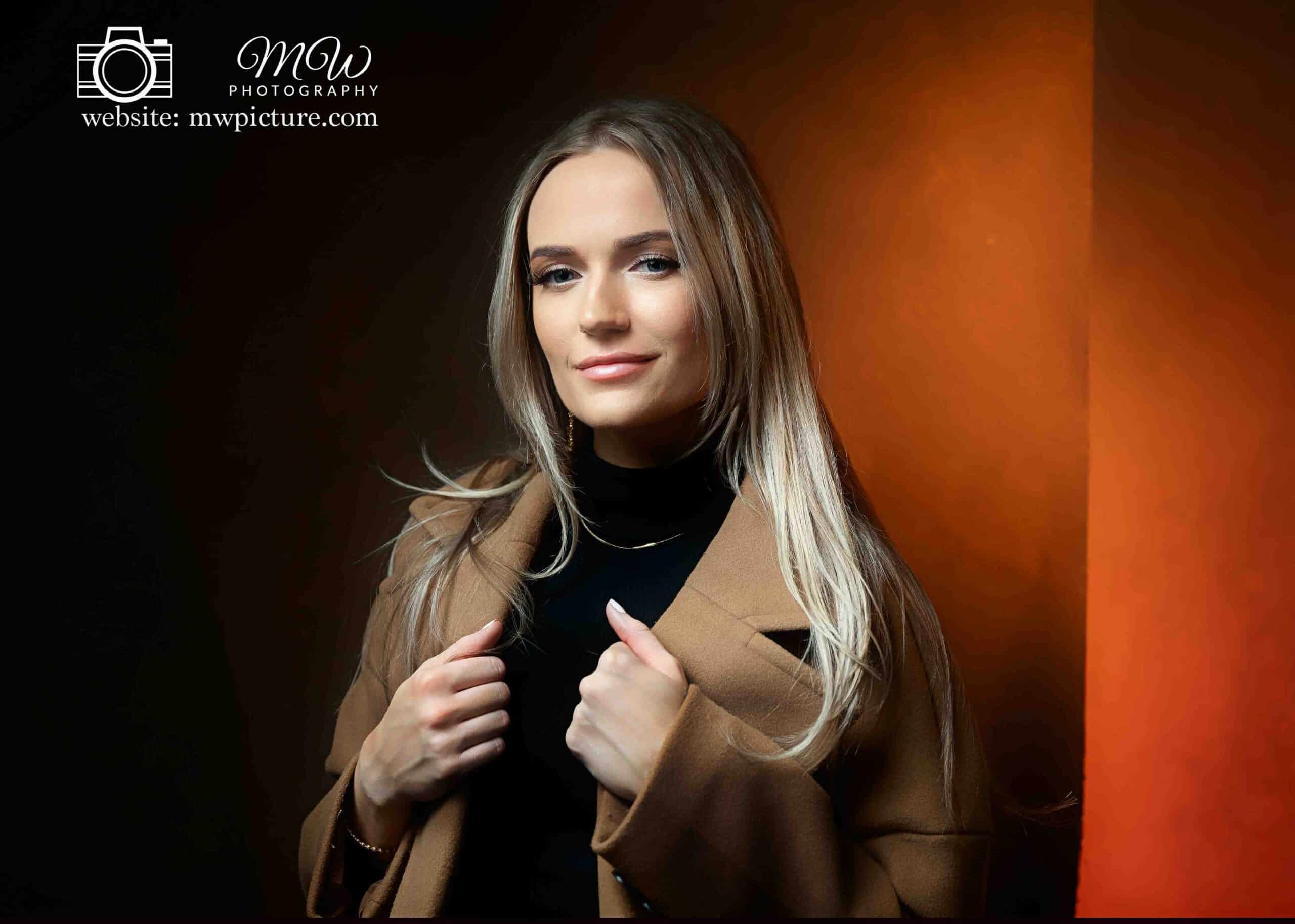 A woman in a black turtleneck and tan coat poses with a slight smile against a dual-tone orange and black background, with a photography logo and website in the corner.