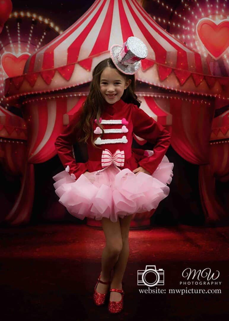 A young girl in a red and pink circus-themed outfit with sequined mouse ears stands smiling against a tent backdrop.