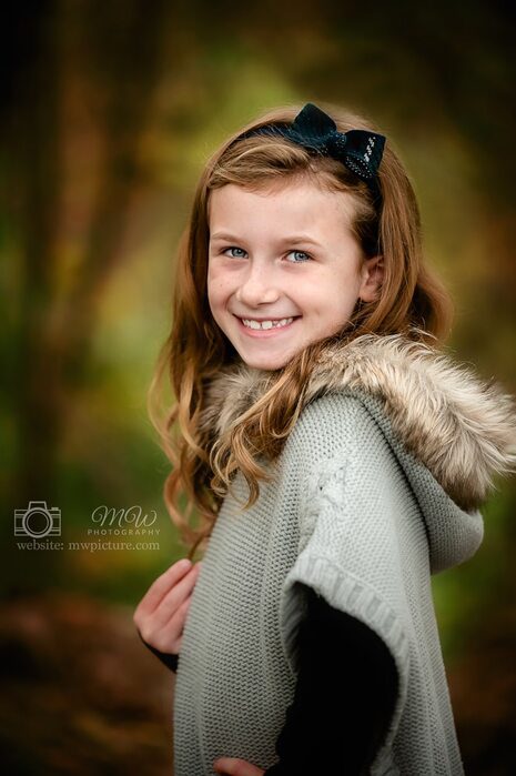 Smiling girl with long brown hair wearing a gray sweater with a fur-lined hood.