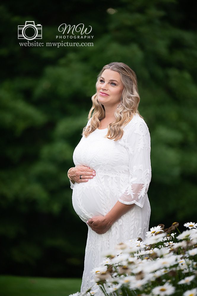 A pregnant woman in white dress standing next to flowers.
