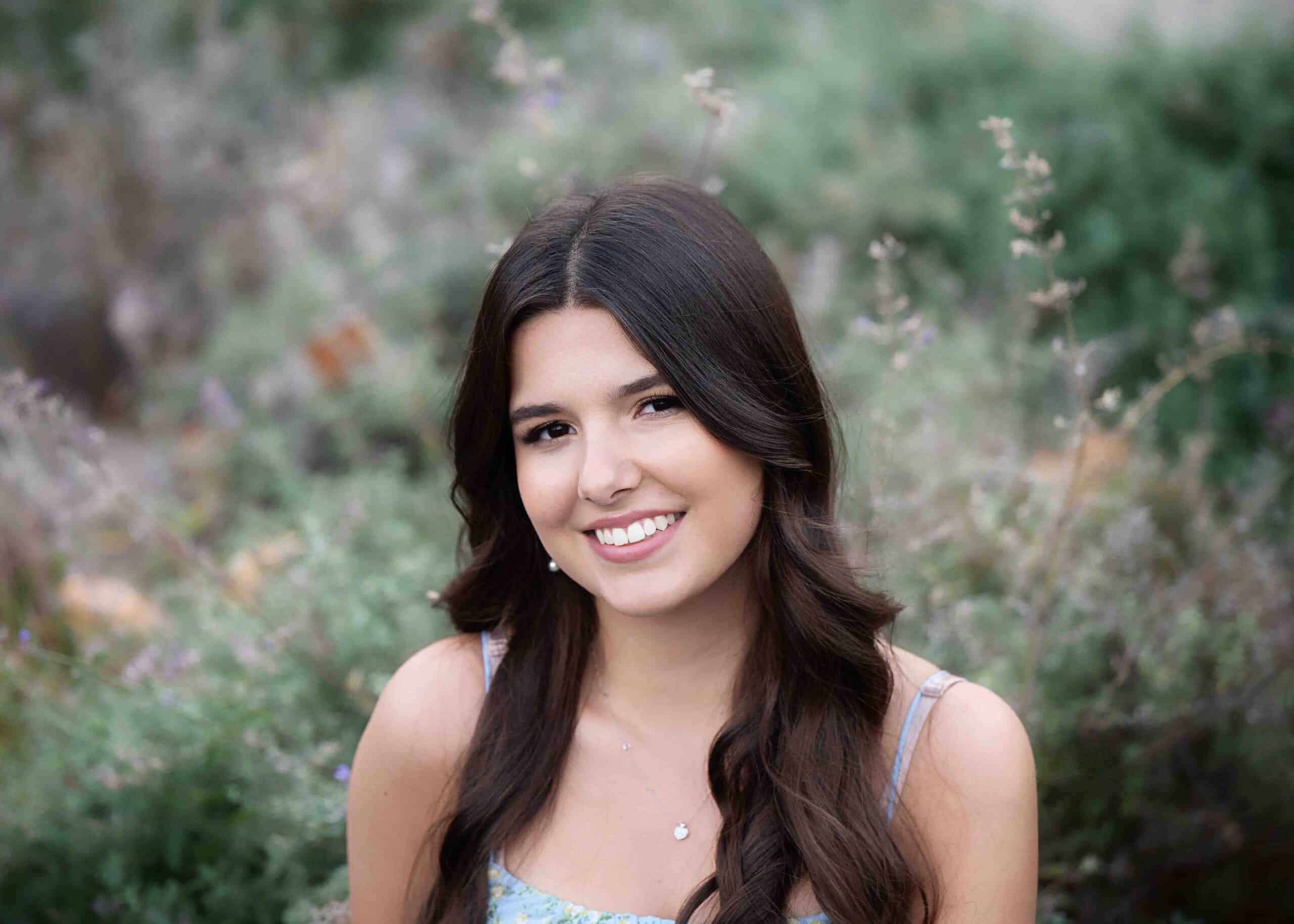 A young woman in a blue dress is smiling in front of bushes.