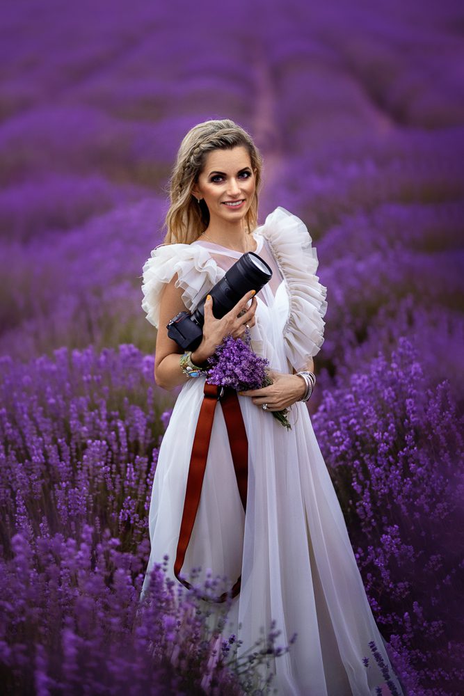 Magdalena Wojton in a Field with Her Camera and a Bunch of Violet Flowers